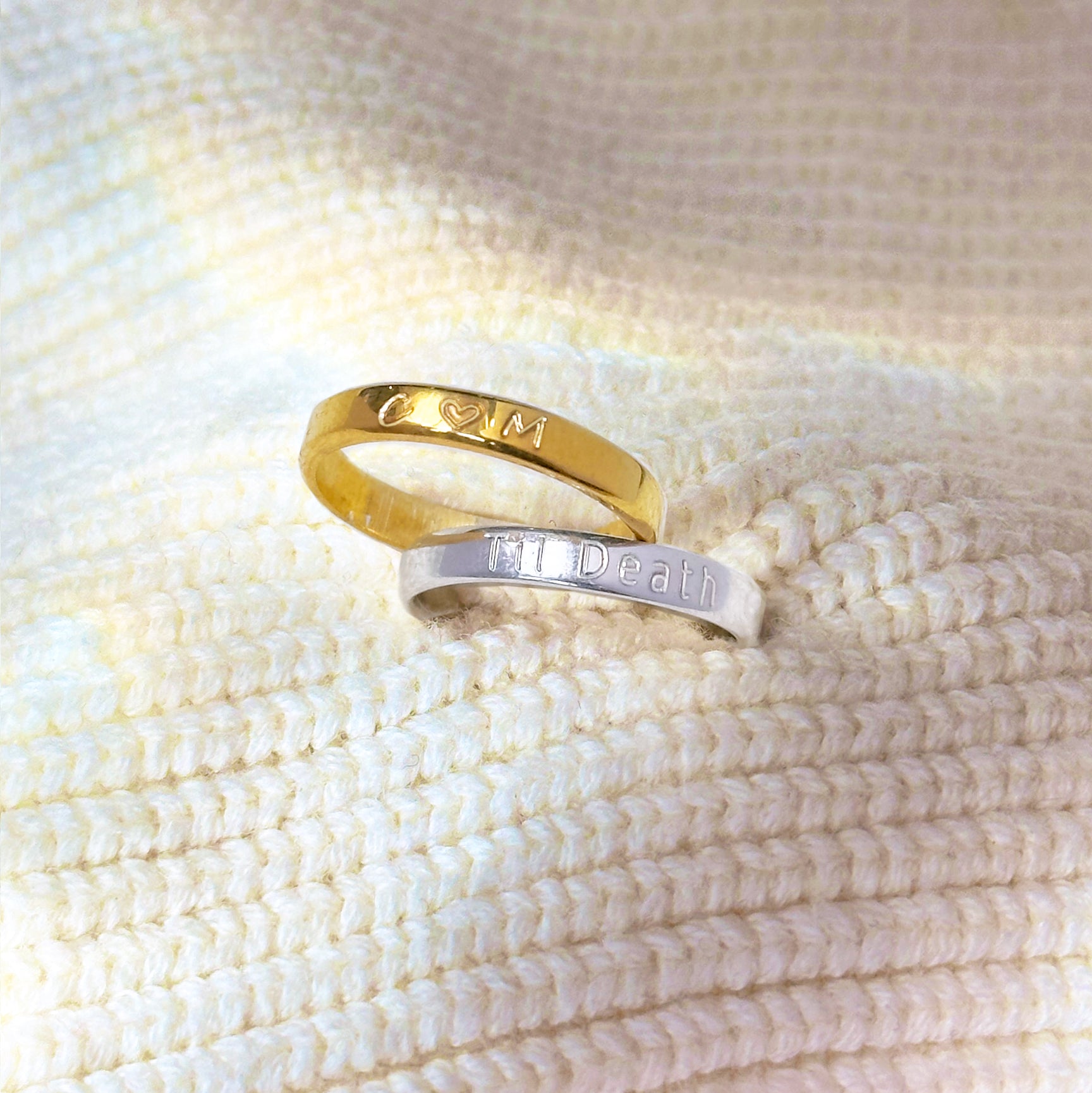 2 Engraved 3mm rings on a white knit background. A gold ring is engraved with initials C (heart) M, a silver ring is engraved with Til Death. Personalized Engraving, Custom engraved rings, dainty stackable name bands for women, monogrammed rings for women, custom message jewelry, secret message jewelry, sterling silver engraved ring bands, unisex jewelry for couples, personalized anniversary rings