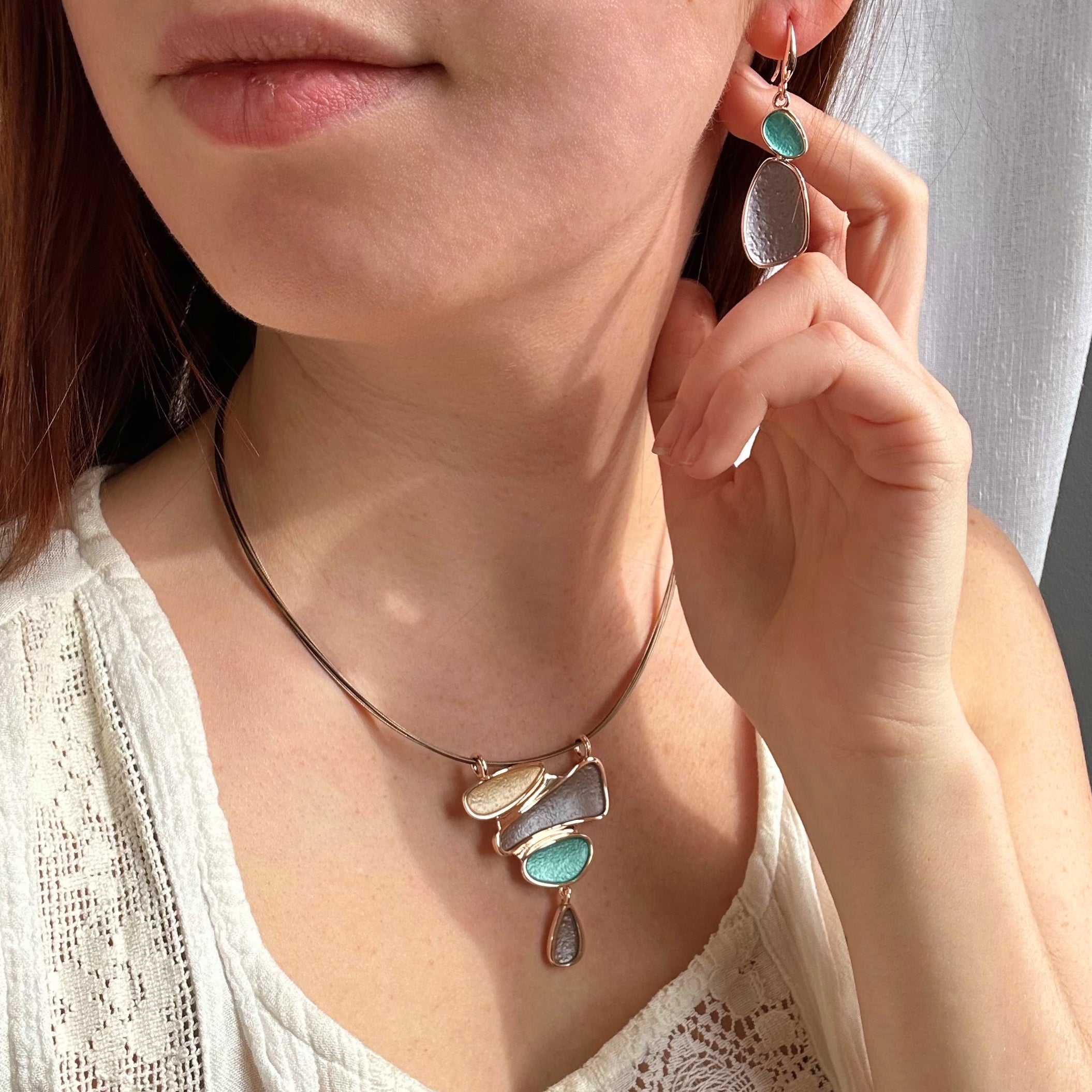 Model wears matching necklace and earring set. Necklace is a pendant with three organic shapes and a dangling teardrop painted in gold, brown, and teal. Necklace has a copper wire chain. abstract stacking stones drop pendant. nature inspired jewelry, geometric jewelry