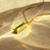 Gold necklace with a green pendant labeled 'SOPHIA' on a textured beige surface.