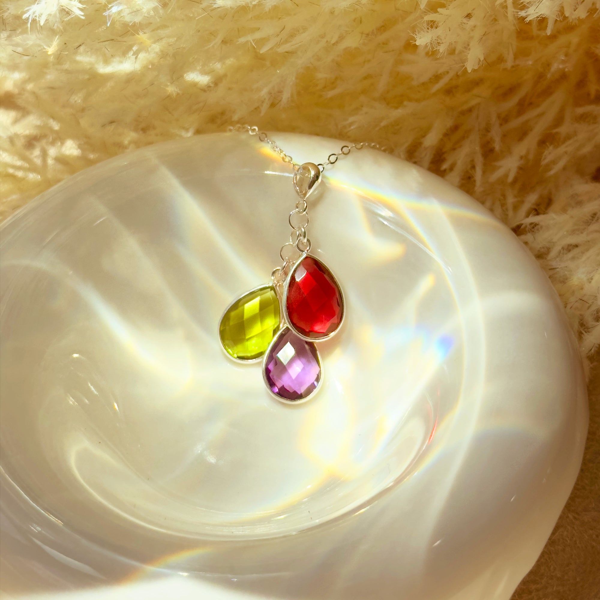 Teardrop birthstone cluster resting on a neutral dish, showing bezel-set stones in vivid colors that represent family months from January to December. A personalized birthstone necklace designed for moms, with options in sterling silver, gold filled, or rose gold filled. Ideal as a sentimental Christmas gift, birthday gift for mom, or push present; easily add stones later for growing families.