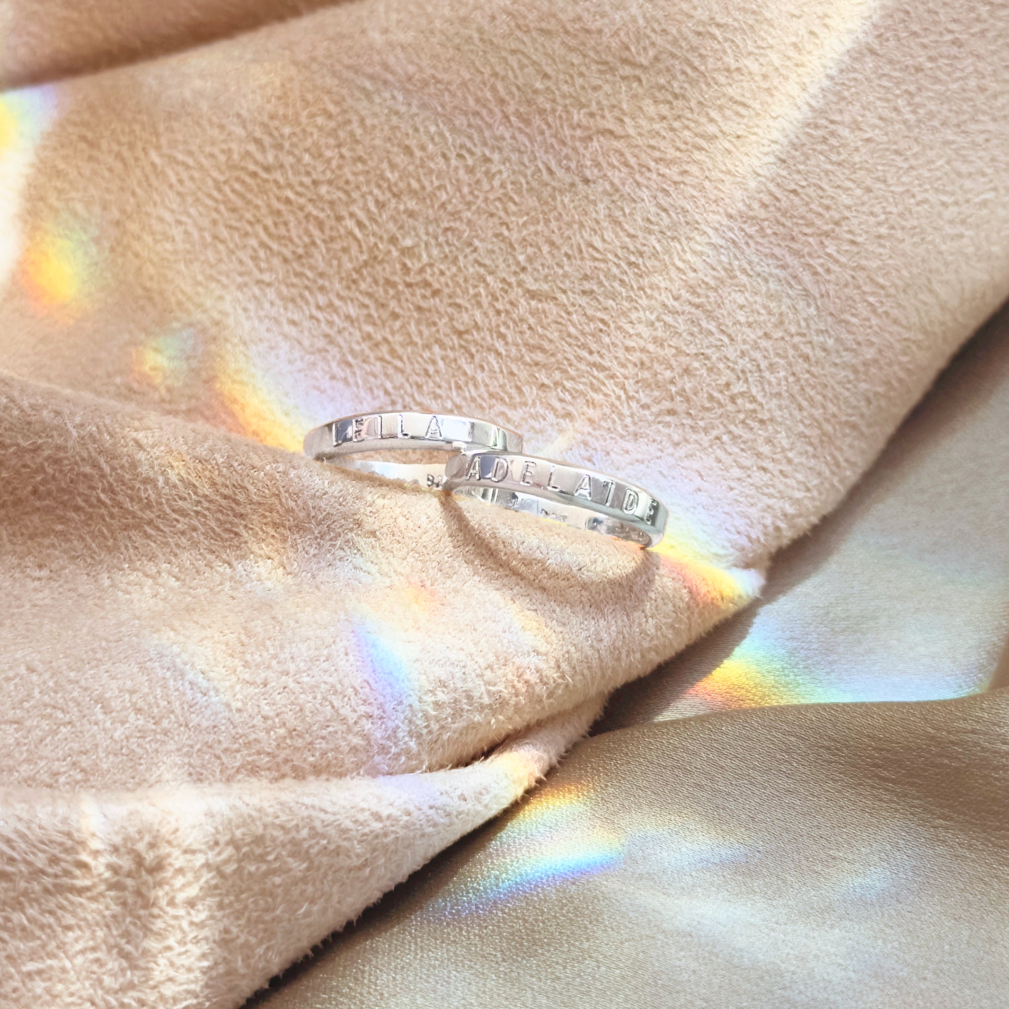 3mm engraved rings in sterling silver shown on soft fabric with rainbow light reflection. Personalized slim bands designed for text engraving including names, dates, and coordinates. Minimalist and unisex, these dainty engraved rings are perfect as stacking rings, promise bands, or anniversary keepsakes. Available in sterling silver, gold, and rose gold, handmade in the USA.