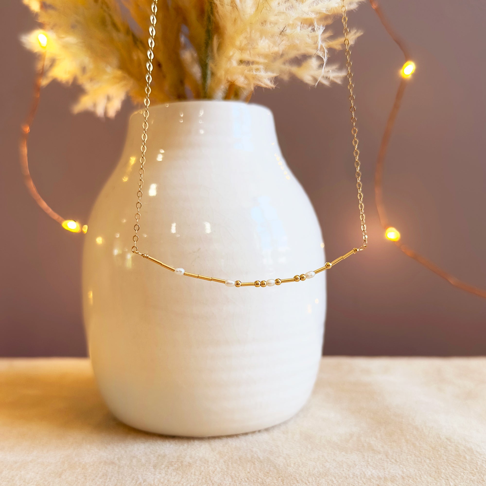 A cozy lifestyle scene featuring the morse code necklace draped on a vase with twinkle lights, highlighting its secret message design. This dainty minimalist necklace is a thoughtful gift for girlfriend, sister, or best friend, and an easy everyday layering piece. Choose sterling silver or gold filled for a hypoallergenic finish; the bead pattern creates subtle personalization without engraving, perfect for birthdays or Christmas gifting.