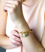 Model is wearing a gold bangle with three name tags and three birthstones including pink chalcedony, moonstone and blue topaz
