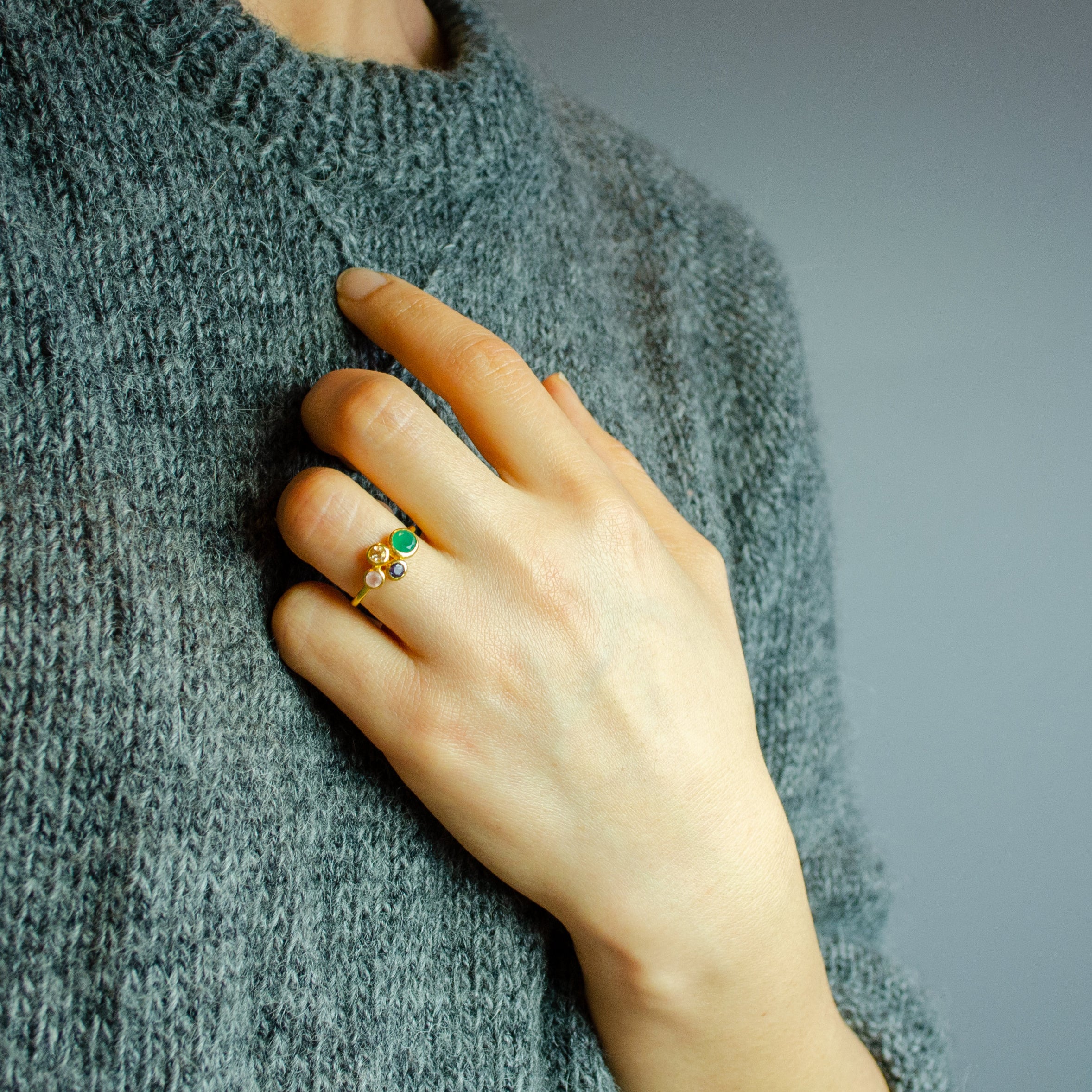 Model in grey top is wearing a single gold plated custom mother of three birthstone ring. Mom stone is green onyx, kid's stones are pink chalcedony, kyanite and citrine on white background