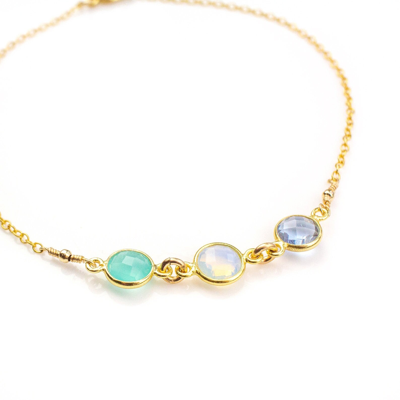 Close-up of a gold birthstone anklet with three bezel-set gemstones in green, opal, and blue tones, highlighting fine craftsmanship and detail. This handmade ankle bracelet can be personalized with any birthstones and is available in sterling silver, gold filled, or rose gold filled. A thoughtful jewelry gift for best friends, matching anklets, or BFF jewelry sets, as well as birthdays, Christmas, or anniversaries. Elegant, dainty, and designed to celebrate love, friendship, and personal connections.