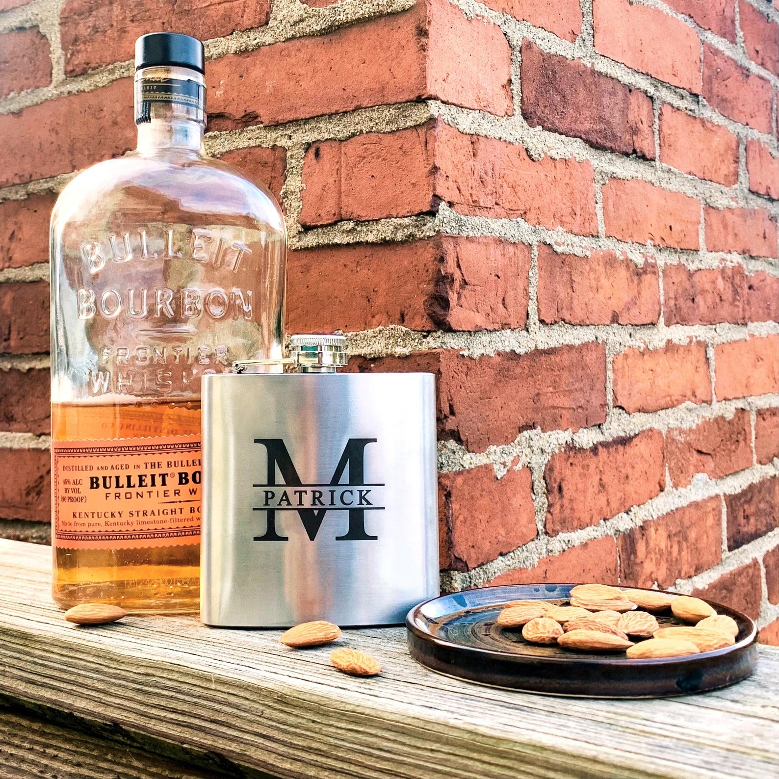 Three custom engraved stainless steel flasks in with different designs: name and message monogram design next to bourbon bottle and almonds. Fantastic personalized gift for party, wedding, christmas, birthday.