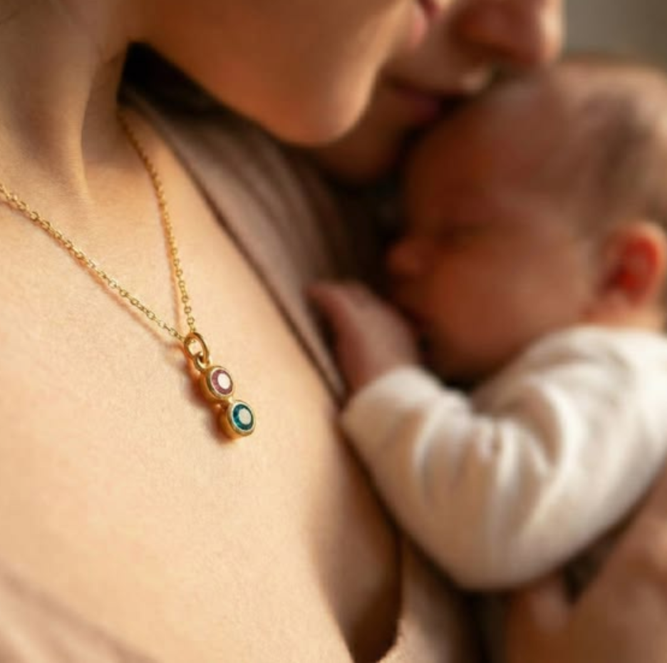 Mother holding a newborn close while wearing a gold birthstone necklace with two colorful stones, styled in a warm intimate portrait that highlights sentimental family jewelry and motherhood.