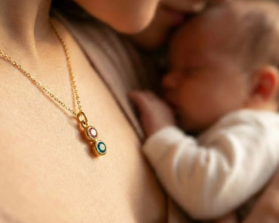 Mother holding a newborn close while wearing a gold birthstone necklace with two colorful stones, styled in a warm intimate portrait that highlights sentimental family jewelry and motherhood.