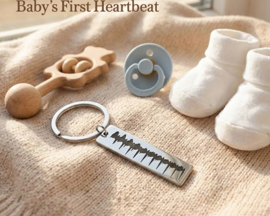 Silver baby heartbeat sonogram keychain on a soft knit blanket beside white baby booties, a pacifier, and wooden rattle, styled in warm sunlight with text reading Baby’s First Heartbeat.