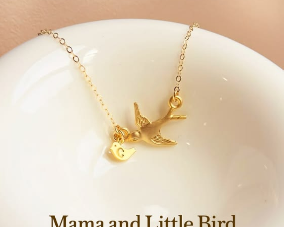 Gold mama and baby bird necklace with a tiny initial charm displayed on a white dish, styled in soft warm light with elegant text, symbolizing motherhood, love, family, and connection.