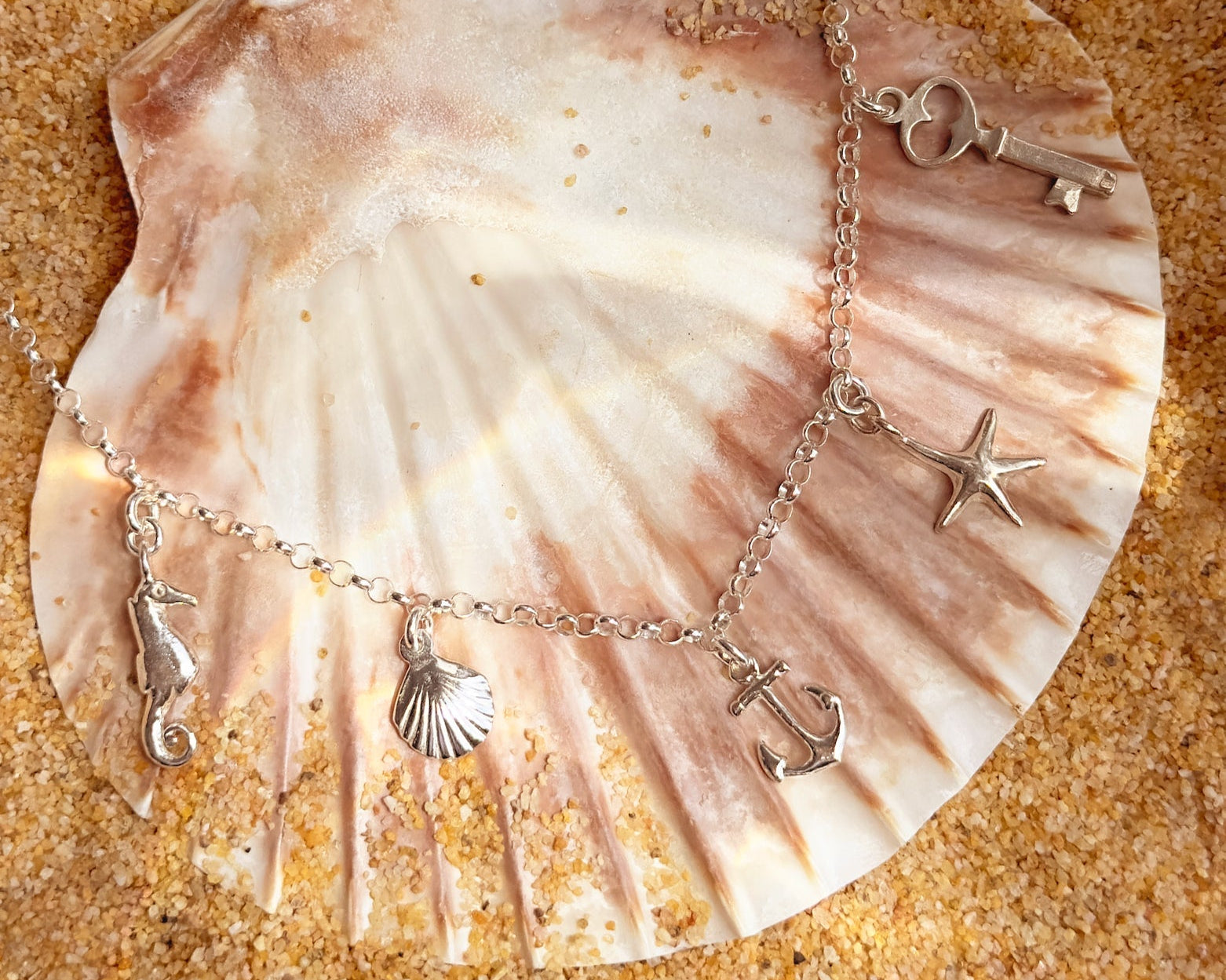 Sterling silver ocean-themed charm anklet with seahorse, seashell, anchor, starfish, key, and heart charms displayed on a scallop shell with beach sand background – perfect personalized summer gift idea.