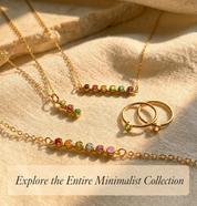 Gold jewelry set with colorful beads on a beige fabric background. The collection features our minimalist birthstone family jewelry collection with necklaces, rings and bracelets.