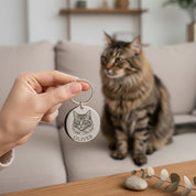 Hand holding a custom engraved pet portrait keychain while a cat sits in the background. Personalized cat portrait keychain featuring detailed engraving from a pet photo. Meaningful pet keepsake gift for cat lovers, designed as a sentimental memorial keychain or everyday accessory honoring a beloved feline.