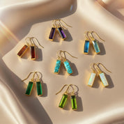 set of baguette cut birthstone drop earrings displayed on soft champagne satin, each pair featuring elongated rectangular gemstones bezel set in warm gold with french hook ear wires, shown in rich purple amethyst, deep garnet red, vivid aqua blue, turquoise matrix, glowing opal white and vibrant green tones, highlighting the modern minimalist design, available in all birthstones for personalized gifting, perfect for birthdays, anniversaries, bridesmaids or everyday statement earrings