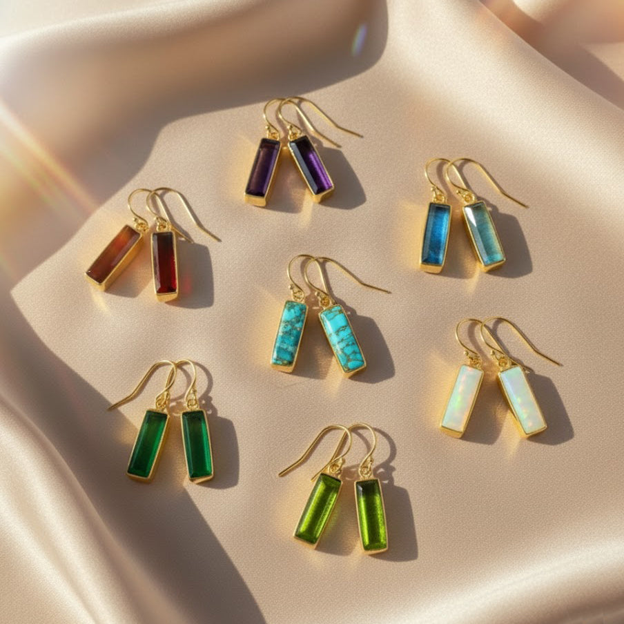 set of baguette cut birthstone drop earrings displayed on soft champagne satin, each pair featuring elongated rectangular gemstones bezel set in warm gold with french hook ear wires, shown in rich purple amethyst, deep garnet red, vivid aqua blue, turquoise matrix, glowing opal white and vibrant green tones, highlighting the modern minimalist design, available in all birthstones for personalized gifting, perfect for birthdays, anniversaries, bridesmaids or everyday statement earrings