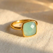 Cushion cut aqua chalcedony ring in polished yellow gold bezel setting resting on neutral linen fabric, featuring a 9mm square pastel blue green gemstone with soft glow, minimalist modern design, dainty gold band, March birthstone jewelry for women, elegant everyday gemstone ring, handcrafted fine jewelry, luxury artisan birthstone ring, stackable gold chalcedony ring gift for her, contemporary bezel set gemstone ring