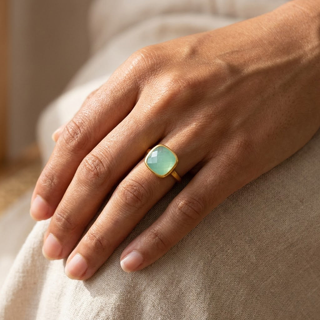 Woman’s hand wearing aqua chalcedony cushion cut ring in polished yellow gold bezel setting, 9mm pastel blue green gemstone, March birthstone ring styled in warm sunlight, minimalist modern fine jewelry, dainty stacking birthstone ring, square chalcedony statement ring for women, handcrafted artisan gold gemstone jewelry gift