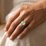 Woman’s hand wearing aqua chalcedony cushion cut ring in polished yellow gold bezel setting, 9mm pastel blue green gemstone, March birthstone ring styled in warm sunlight, minimalist modern fine jewelry, dainty stacking birthstone ring, square chalcedony statement ring for women, handcrafted artisan gold gemstone jewelry gift