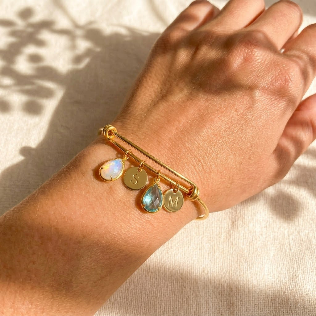 Custom family birthstone bangle bracelet on wrist with teardrop stones and initial charms in gold.