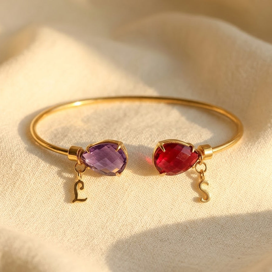 Two birthstone initial bangle bracelet in gold with purple and red teardrop gemstones and matching letter charms, personalized for two loved ones.