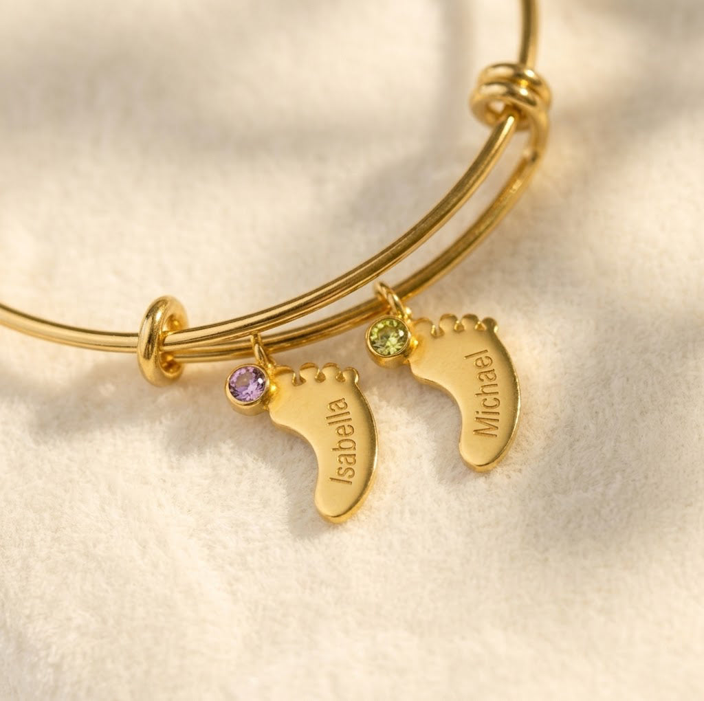Gold custom mom bangle bracelet with engraved footprint charms, children’s names, and tiny faceted birthstones on a warm neutral background.