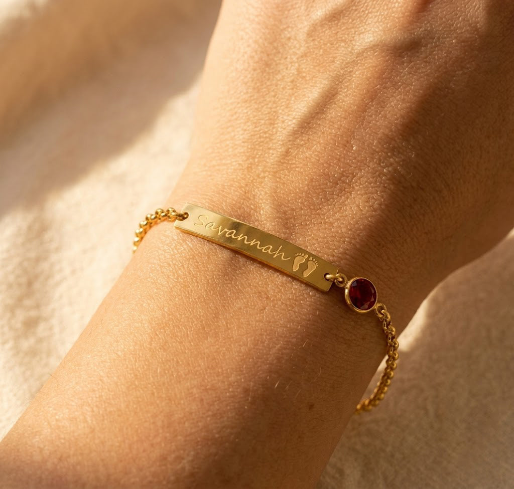 Dainty personalized baby footprint birthstone bracelet in yellow gold filled, styled on wrist. Features custom engraving and birthstone charm. Ideal push present, Mother’s Day jewelry, or new mom gift. Handcrafted adjustable bracelet in sterling silver, gold, or rose gold.