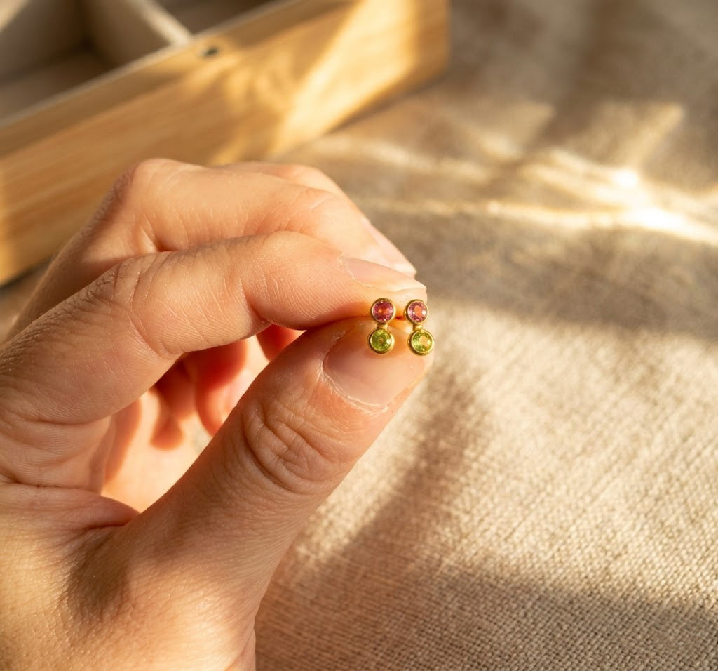 Close-up of hand holding colorful custom birthstone bar studs, showing tiny personalized gemstone earrings made in sterling silver or 18K gold.