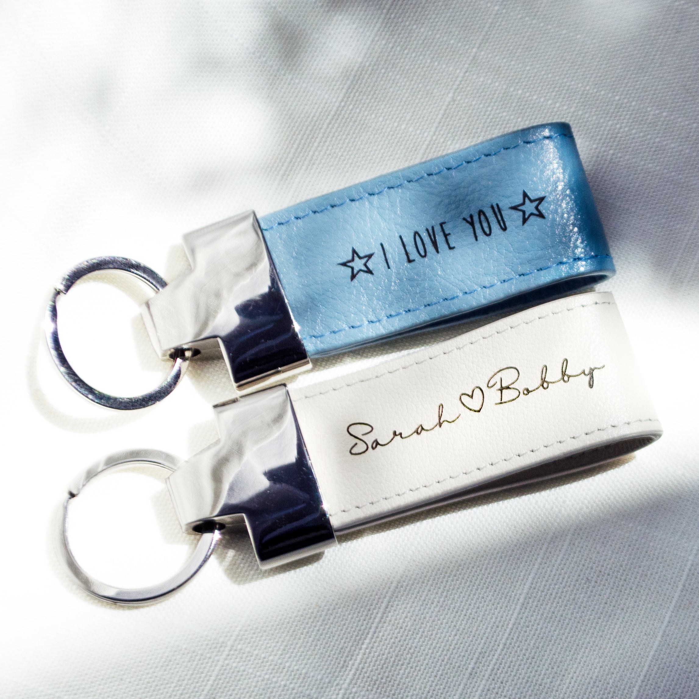 Two keychains at a diagonal angle rest on a textured cloth background. A silver blue keychain says I Love You in Sunshine font with stars on each side. A white keychain says Sarah (heart) Bobby in Grace font.Leather Keychain White Blue Magenta Brown Zinc Alloy Metal Keyrings Personalized Gift Personalized Key Fob College Dorm Gift College Grad Gift Graduation Gift for Him or Her Natural Leather Custom Gift for Women, leather anniversary gift, leather wedding anniversary gift, 3rd anniversary gift