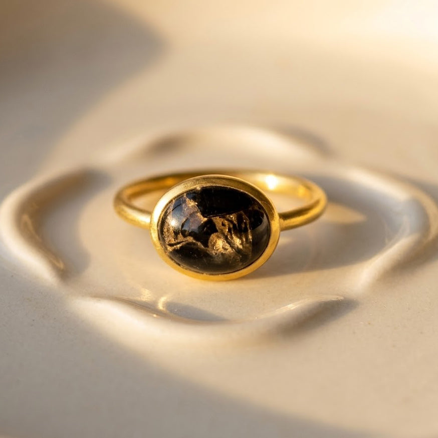 Oval black obsidian cabochon ring in yellow gold shown on warm beige background