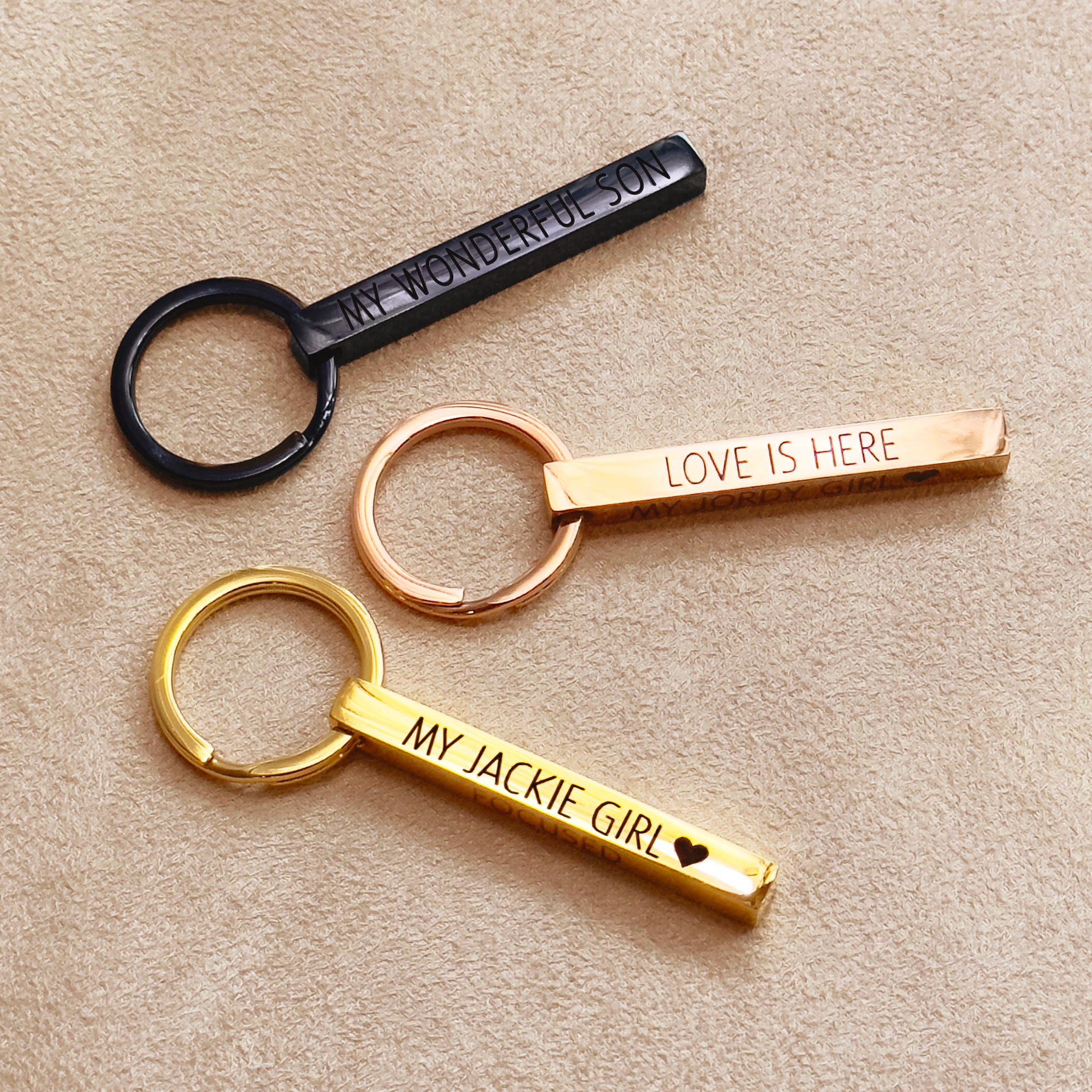Trio of engraved bar keychains—matte black, classic gold, and rose gold—styled on soft tan fabric. Each features different engraving lengths to demonstrate personalization capacity across one to four sides. Ideal for initial keychain sets, travel coordinates keychain, or motivational quote gifts for graduates and new jobs.