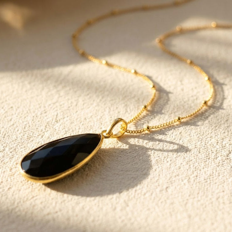 Large faceted Black Onyx teardrop pendant necklace in yellow gold displayed in warm natural light.
