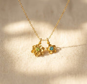 Gold birthstone flower charm necklace on warm beige fabric with colorful gemstone centers, meaningful personalized necklace for mom.