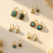 Labradorite earrings in multiple styles arranged in warm sunlight, showing blue-green flash gemstones in gold settings, including teardrop and round drop earrings for everyday wear or gifting.