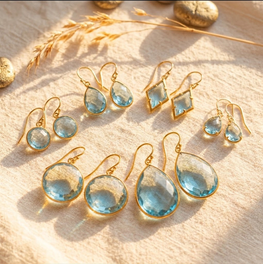 Faceted blue topaz earrings in seven teardrop, round, and geometric styles on cream fabric.