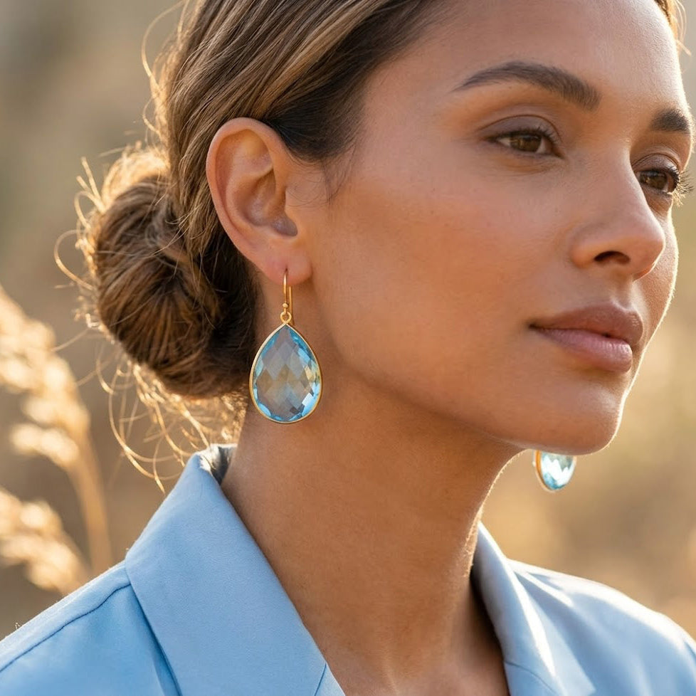 Woman wearing large teardrop blue topaz earrings with gold ear wires in natural sunlight.