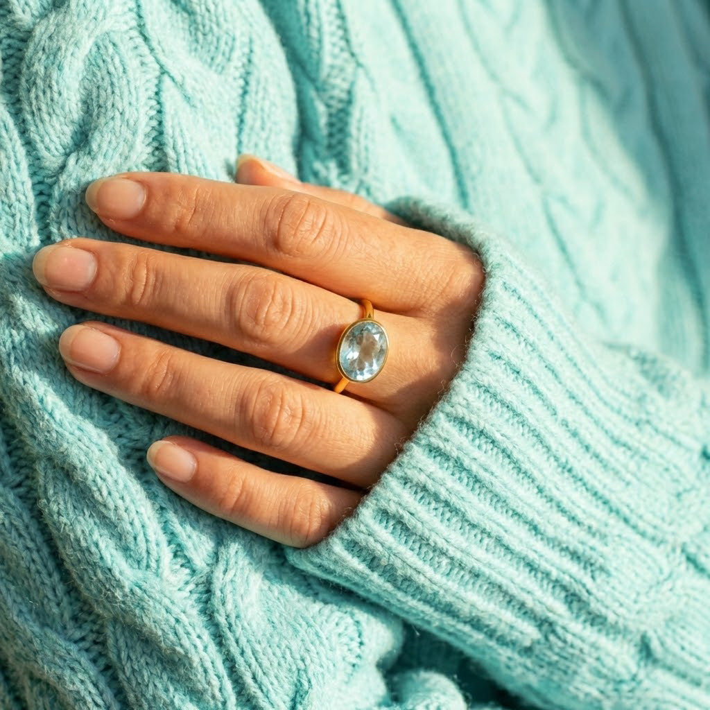 Blue topaz December birthstone ring worn on hand with cozy knit sweater, oval gold gemstone ring for women, minimalist stacking jewelry for everyday elegance