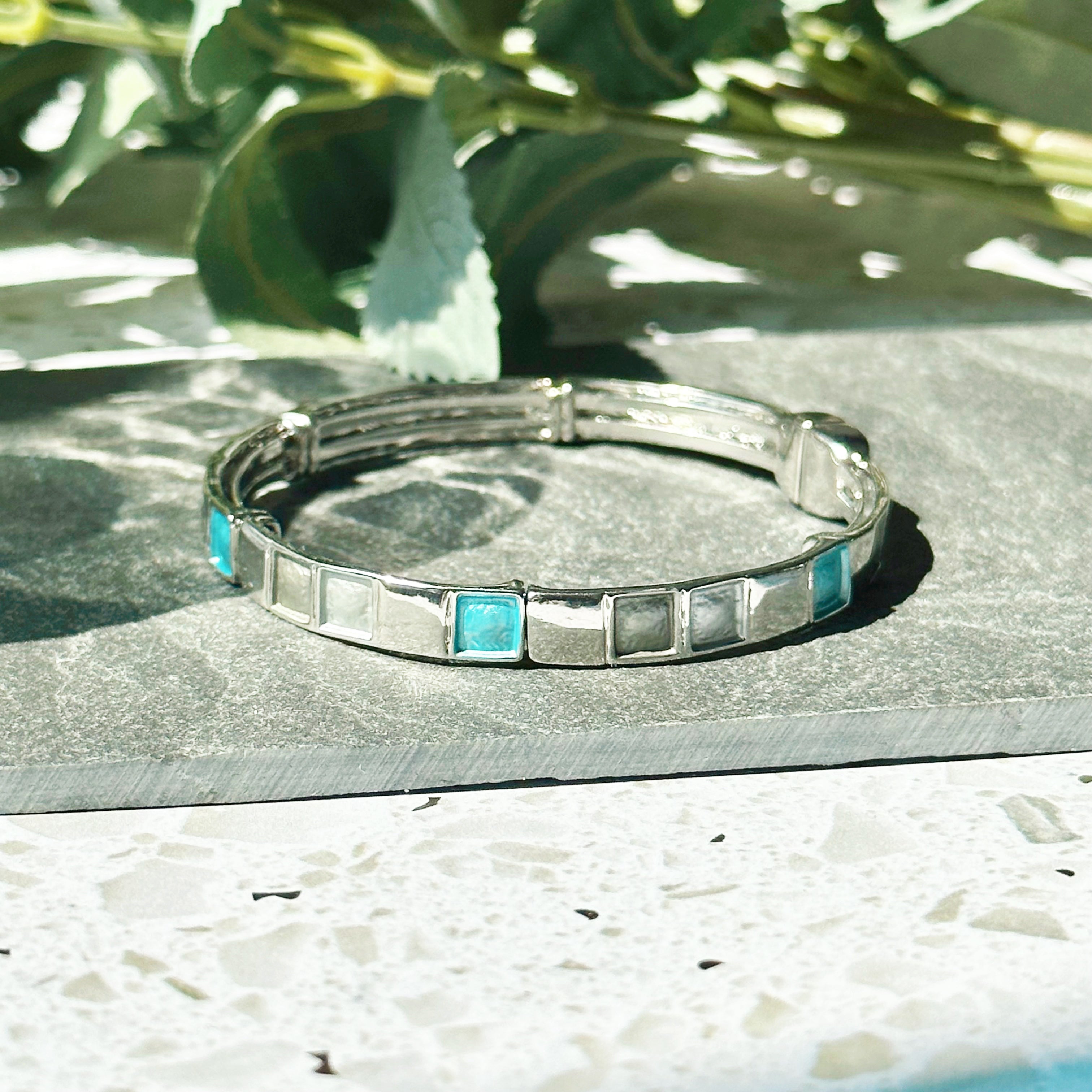 Rhodium plated cuff bracelet with a magnetic closure with square indents painted in silver, gray, and teal enamel is placed on a black stone tile with shadows cast by a glass and a leafy plant