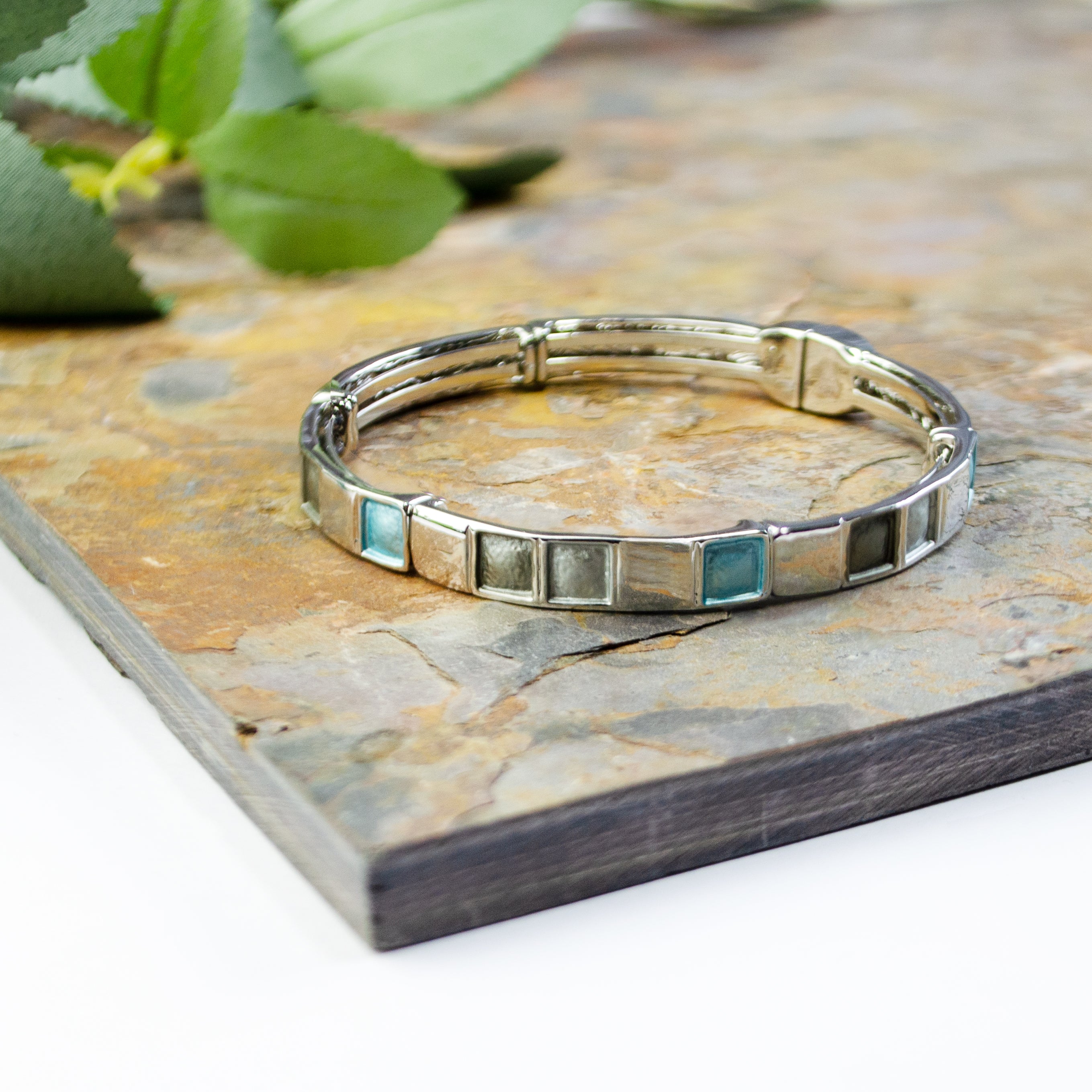 Rhodium plated cuff bracelet with a magnetic closure with square indents painted in silver, gray, and teal enamel placed on a brown and gray textured stone tile