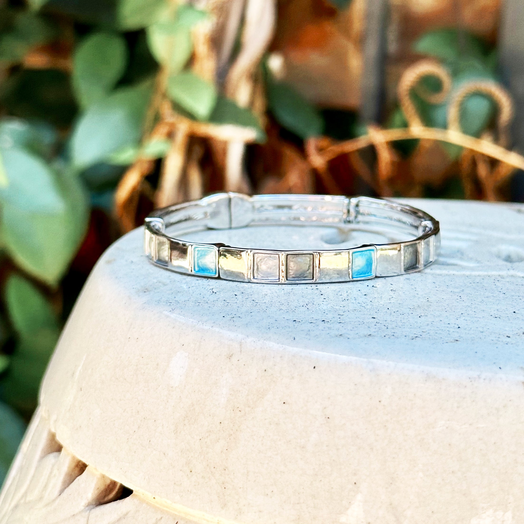 Rhodium plated cuff bracelet with a magnetic closure with square indents painted in silver, gray, and teal enamel placed on a ceramic pot