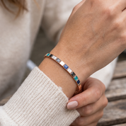 Delicate rose gold bangle bracelet with multicolor square accents in turquoise, blue, white, gray and black, shown on a woman’s wrist.