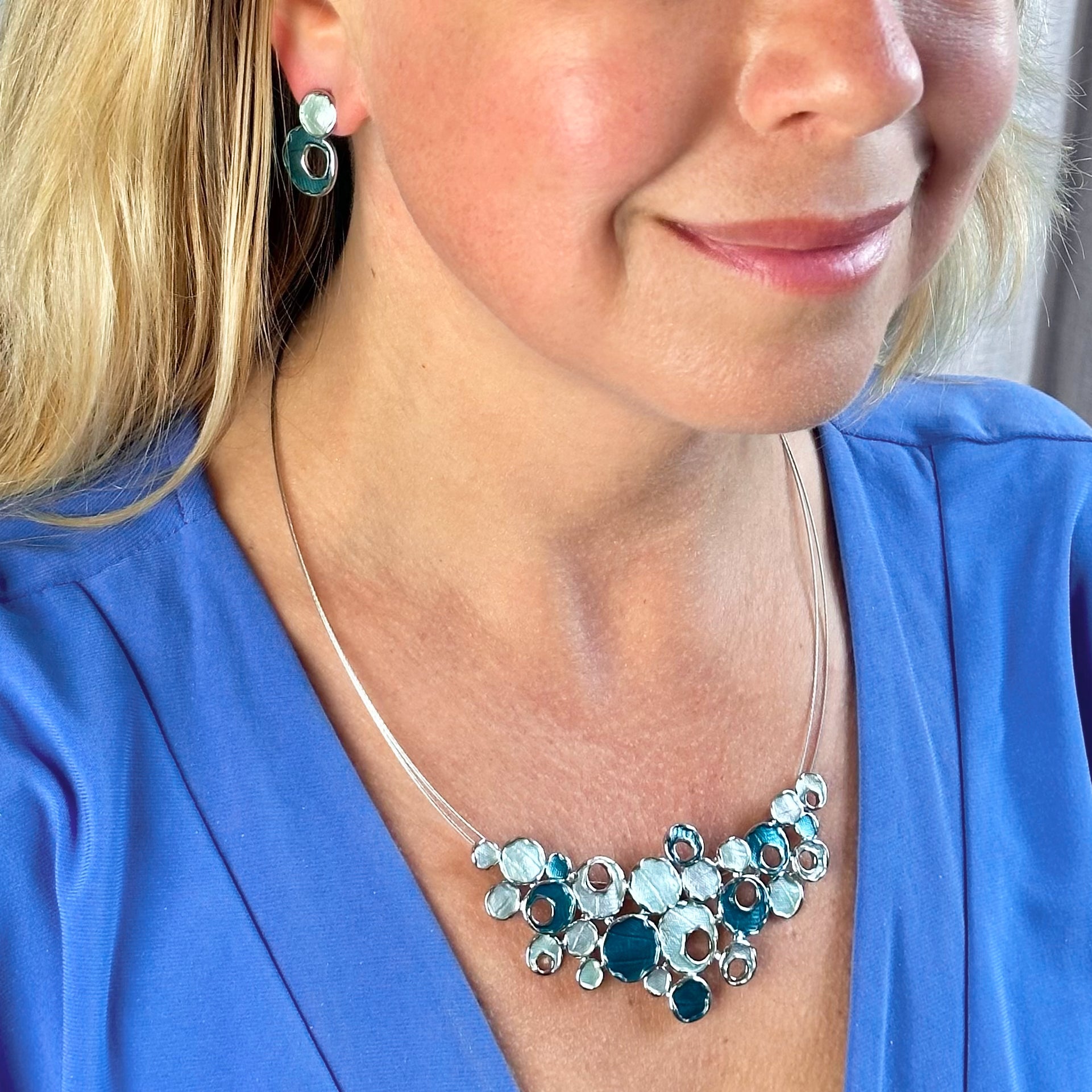 Matching Earrings and Necklace worn by a model with a steel cable chain with an organic triangular shaped pendant made up of rhodium plated circles painted in blue and rose enamel. Some of the circles contain a circular cut out. Boho jewelry, geometric necklace, geometric jewelry