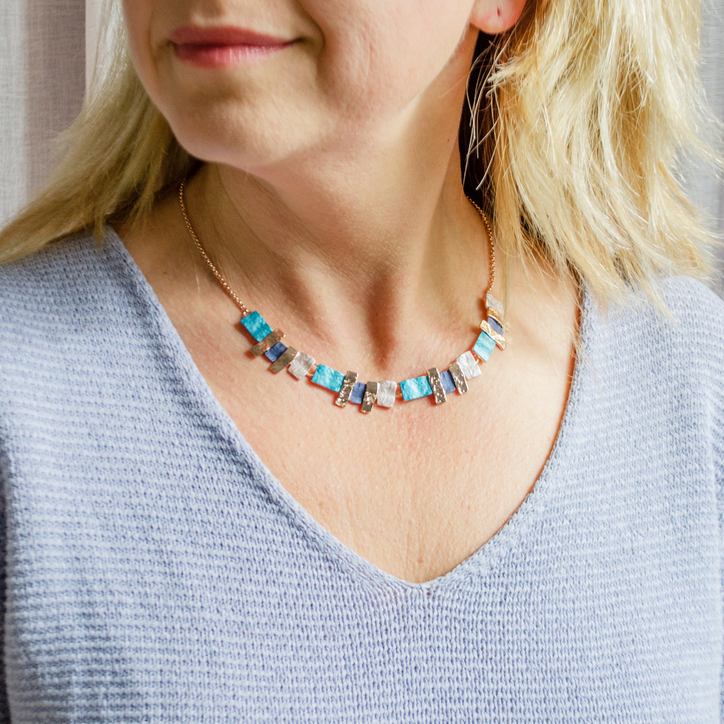 Pair of rose gold plated textured stacked squares necklace with rose gold, purple and pink square and rectangle pendants worn by a blonde model wearing a blue blouse