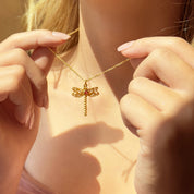 Dainty gold dragonfly necklace with intricate filigree wings and a tiny red birthstone held between a woman’s hands, feminine nature-inspired jewelry in warm sunlight; delicate insect pendant on fine chain, whimsical minimalist necklace gift for her, boho summer accessory.