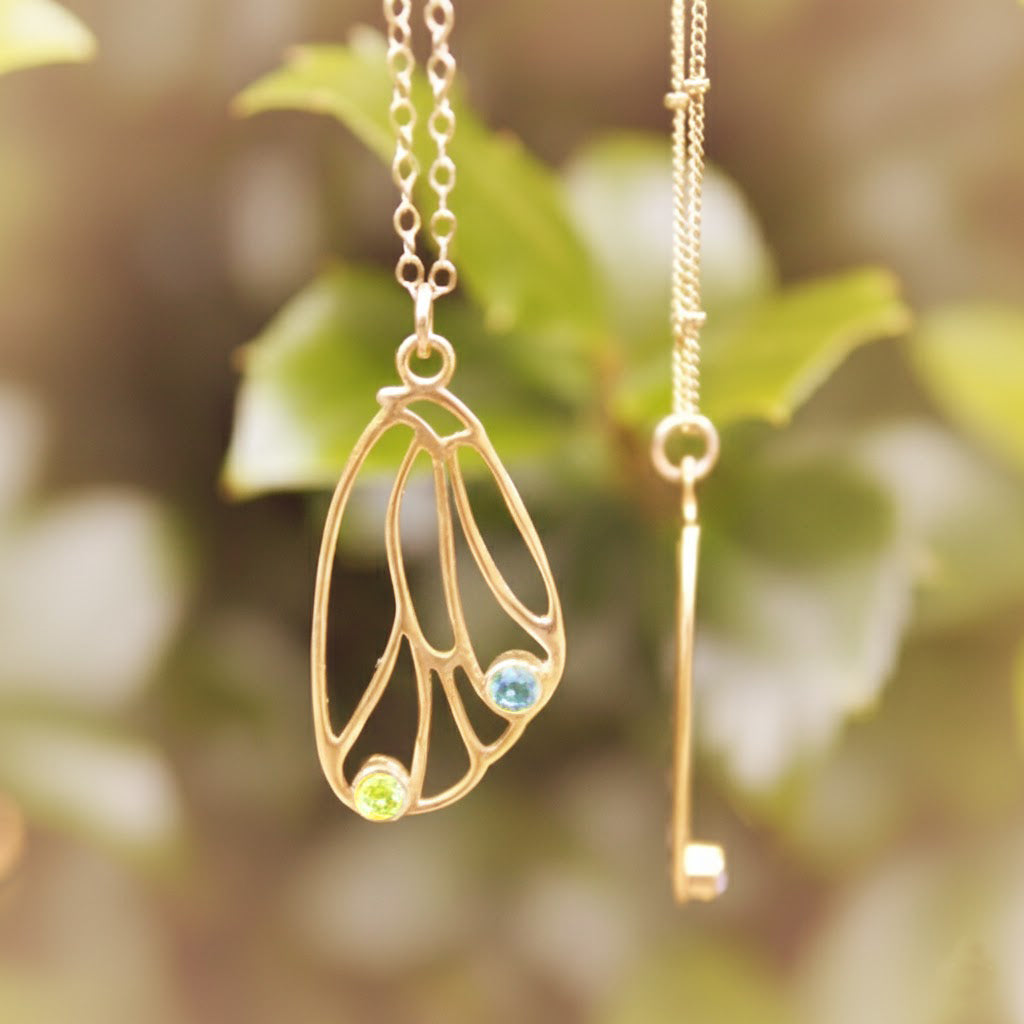 Gold butterfly wing necklace with blue birthstone and key charm hanging from branch, symbolic custom birthstone jewelry for nature and butterfly lovers