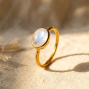 Oval rainbow moonstone ring in yellow gold vermeil with a glowing blue flash on a warm neutral background.