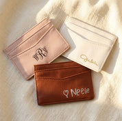 Personalized PU leather card holders in pink, white, and brown with engraved names and monograms on a warm neutral background.