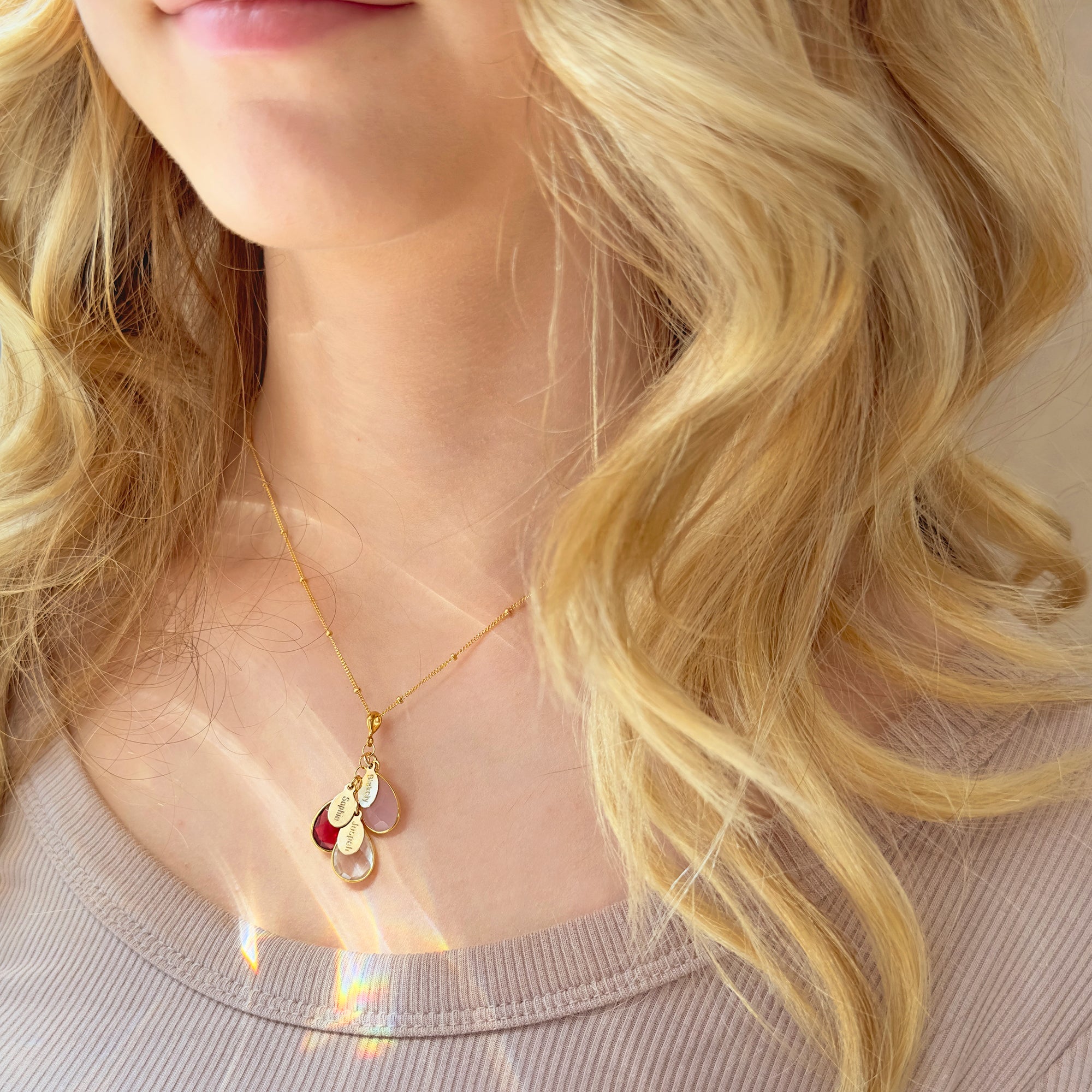 A person in a beige shirt wears a cascading necklace with birthstones and name tags visible. The necklace features the October birthstone, the July birthstone and the April birthstone. 