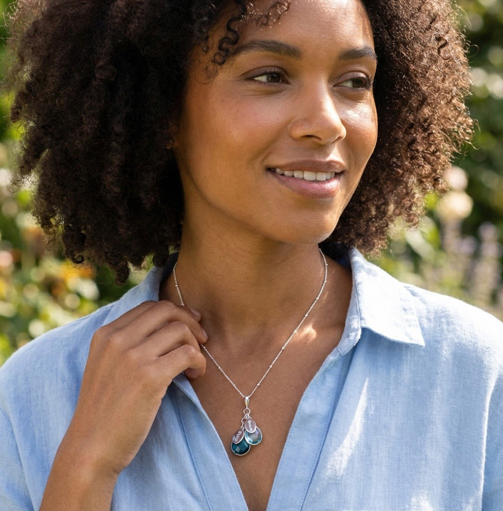 A mom in a white sweater wears a cascading necklace with birthstones and name tags visible. The necklace features the February birthstone, the December birthstone and the September Birthstpne birthstone: Blue Topaz, Kyanite and Purple Amethyst.
