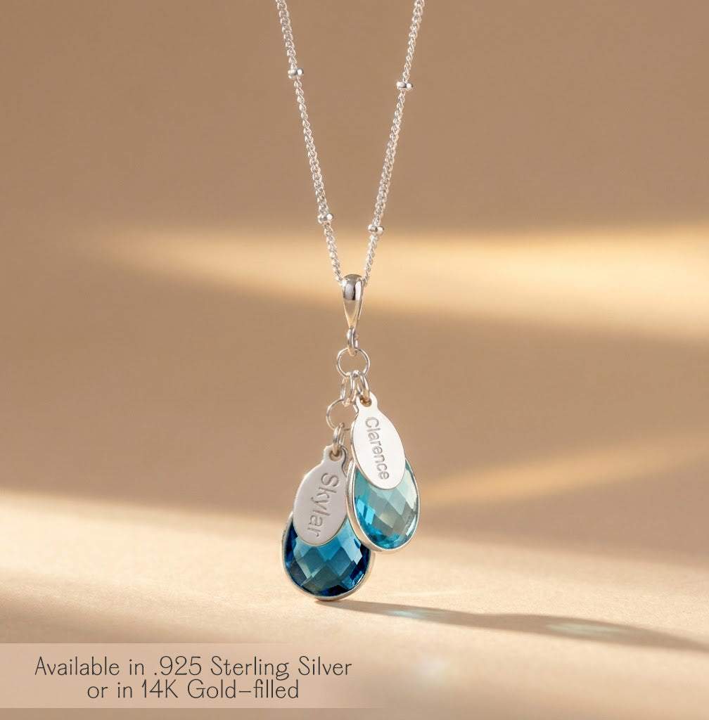 Sterling silver custom birthstone mom necklace with two faceted teardrop aqua blue stones and matching oval name charms engraved with children’s names, hanging from a delicate satellite chain on a clean white background. Personalized family necklace for moms, new moms or grandmas, beautifully showcasing kids’ birth months, a meaningful Mother’s Day, birthday or Christmas gift idea and everyday keepsake jewelry piece.