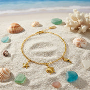 Custom charm anklet flat lay styled with seashells and natural stones, gold filled personalized anklet with multiple dangling charms, handmade summer anklet inspired by beach jewelry aesthetics, minimalist ankle bracelet for women designed for layering and warm weather outfits, adjustable charm anklet crafted with attention to detail and meaningful charm selection